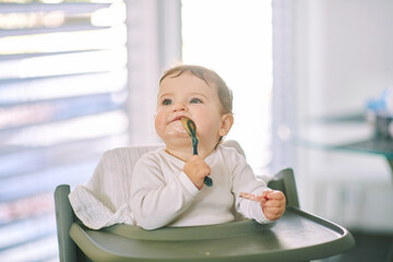 Portrait of adorable messy baby sitting in high chair, holding spoon