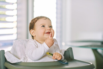 Portrait of adorable messy baby sitting in high chair, holding spoon