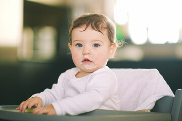 Portrait of adorable messy baby sitting in high chair, holding spoon