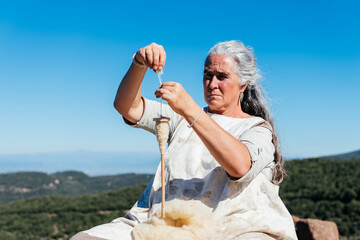 Senior woman making thread with thread maker spinning wheel against mountain and blue sky