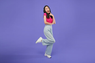 full body of a thoughtful Asian woman who keeps her hand on her chin and looks pensively above poses against a purple background blank copy space for your advertising content thinks about future