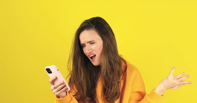Mad and shock young brunette beautiful woman using mobile phone isolated on yellow background. Yelling unhappy woman hold mobile phone. Disappointed sad upset lady horrified impressed news. WTF. Oh no