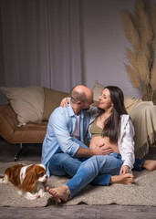 A man sits on the floor, a pregnant woman with a bare belly sits on the sofa. He holds small socks in his hands, and a dog sits next to them. Cavalier King Charles Cocker Spaniel