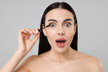 Emotional young woman applying mascara on grey background