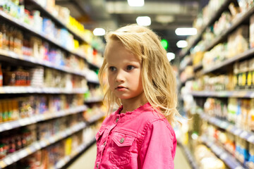 Child girl 5 year old at shelves with groceries in store background, bored looking away. Portrait of cute kid girl shopping choice buying in supermarket. Retail shopping concept. Copy ad text space