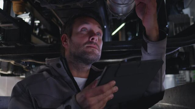 Auto mechanic examining truck part underneath and using digital tablet. Repair service garage worker checking pipe under lifted trailer