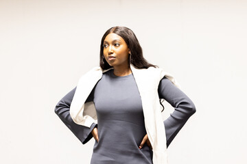 The image presents a determined and confident young businesswoman of African descent standing with her hands on her hips, dressed in a chic, form-fitting grey dress with a casual white jacket over her
