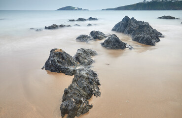 Shoreline boulders buried in sand face an advancing tide at dusk, looking from Bantham beach towards Burgh Island, on the south coast of Devon, England