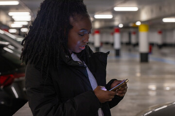 The image portrays a young woman engaged with her smartphone in a dimly lit parking garage. Her...
