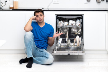 Tired and bored man putting dirty plates in dishwasher machine in the kitchen. Household and...