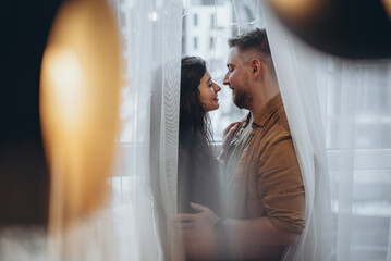 A young couple stands near the window, hugging and looking at each other, wrapped in curtains. The city is blurry in the background. In the foreground is a chandelier with a light bulb and glare - obrazy, fototapety, plakaty