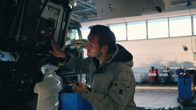 Auto mechanic examining truck under hood and using digital tablet in sunlit workshop, tracking shot. Repairman walking in repair service garage, inspecting trailer vehicle