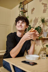 Fototapeta na wymiar happy and curly young man holding organic tofu burger and smiling in plant filled vegan cafe