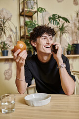 Fototapeta na wymiar positive young curly man holding plant-based tofu burger and talking on smartphone in vegan cafe