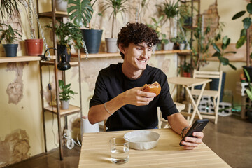 Fototapeta na wymiar positive young curly man holding plant-based tofu burger and using smartphone in vegan cafe