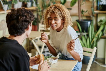 excited african american girl chatting with curly boyfriend near plant based meal in vegan cafe