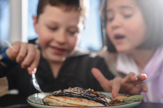 Two children enjoying a meal together, with a boy holding a fork over a pancake topped with syrup and a girl pointing towards it