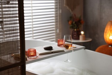 White wooden tray with glass of rose wine, book and burning candles on bathtub in bathroom