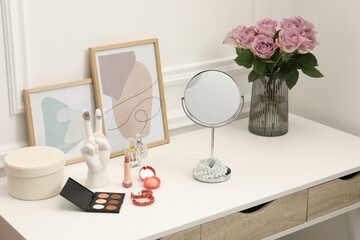 Mirror, cosmetic products, perfumes and vase with pink roses on white dressing table in makeup room
