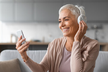 senior lady in headphones listening to music using smartphone indoors