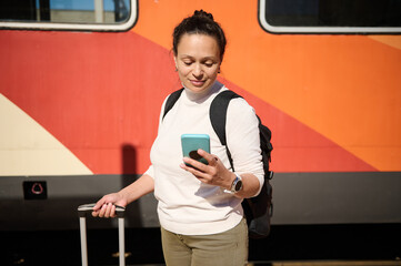 Young woman checking mobile app on smartphone, standing at railway platform, waiting to board the...