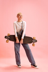 Full length portrait of young attractive woman in denim overall posing holding skate board against...
