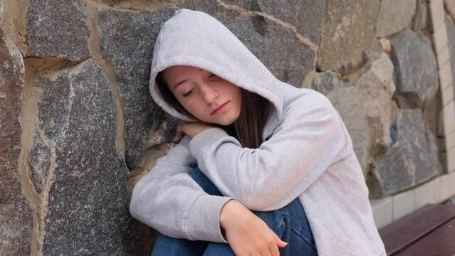 Sad depressed teenage girl in a hood sleeps alone on the street, feeling angry and lonely. Growing crisis and anxiety. Concept of self-acceptance and self-analysis. Homeless