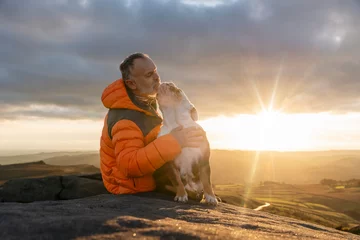 Fotobehang old mature man hiking in mountains with his dog, exercise and fitness for wellness, healthy lifestyle and smile. Face of a senior mature gentleman with bulldog sitting on a rock, enjoying a calm day © Iryna
