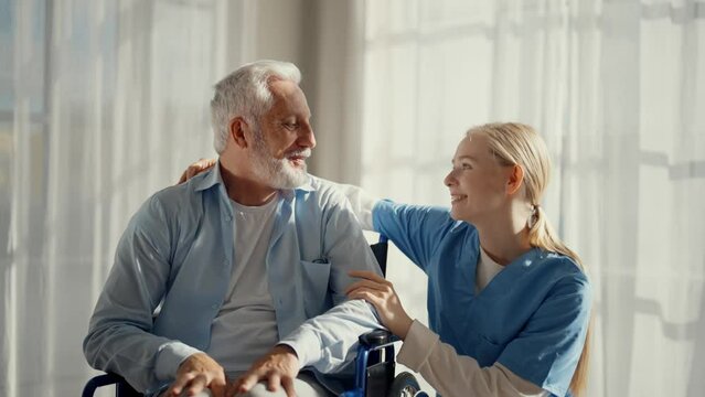 Nurse talking senior man in wheelchair. Female social welfare physical therapist looks after elderly male, listening, providing medical care at home. Living with legs paralysis, special needs people.