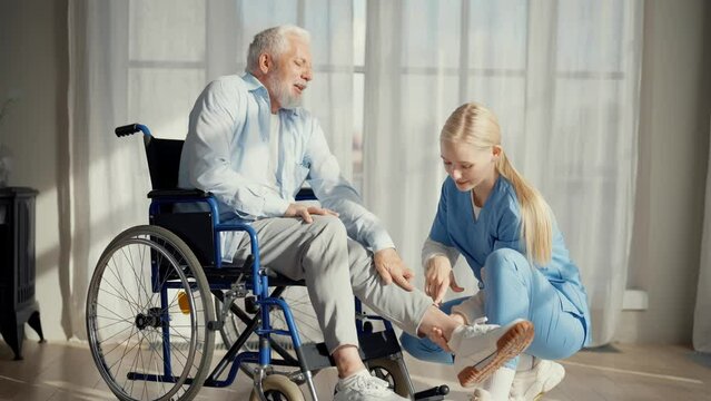 Nurse talking senior man in wheelchair. Female social welfare physical therapist looks after senior male, listening, providing medical care in clinic. Living with legs paralysis, special needs people.