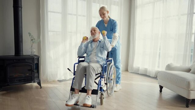Nurse woman cares old man in wheelchair. Paralyzed elderly man does exercises under supervision of a nurse supporting at home. Living with spine legs paralysis, special needs people health concept.