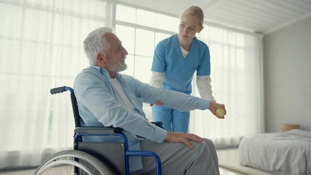Nurse woman cares old man in wheelchair. Paralyzed elderly man does exercises under supervision of a nurse supporting in clinic. Living with spine legs paralysis, special needs people health concept.
