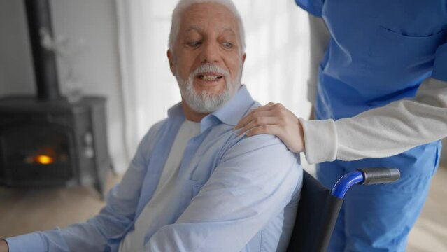 Old man in wheelchair talks with nurse. Senior paralyzed male thanks social worker woman for help support at home. Caregiver holds hand on patient shoulder stands near. Legs paralysis, special needs.