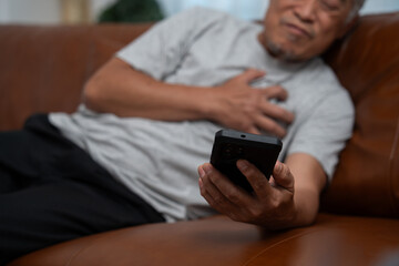 Senior male asian presses hand to his chest has a heart attack suffers from unbearable pain in the living room, lonely men try using the smartphone to call for Emergency help.