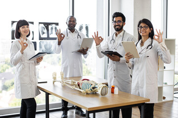 Fototapeta na wymiar Young multiethnic medical students on anatomy lesson in modern classroom showing sign ok. Middle aged male doctor professor teaching anatomy using human skeleton model with with body organs.