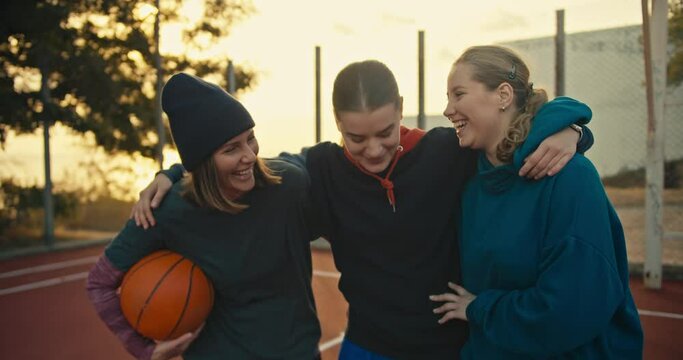 Close up a trio of blonde girls in sportswear with a basketball on a red summer court on the street stand hugging and talking after their match in the morning at sunrise