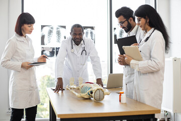Fototapeta premium Human skeleton model on the table in classroom. Group of international medical students with male senior teacher standing near table and having anatomy lesson.