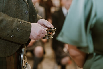 close up of a man holding silver quaich in his hands beside a bridesmaid in green dress at a...