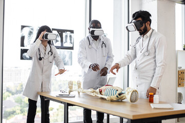 Education, science, vr, medical and health concept. Group of multiethnic doctors, biologists, archeologists, scientists wearing VR goggles study human skeleton in modern laboratory. - obrazy, fototapety, plakaty