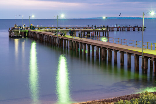 Fototapeta Fishermen and tourists on a wooden pier over a calm coastal bay at twilight