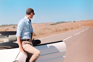 Handsome young man in sunglasses leaning on luxury white convertible while standing stop on road