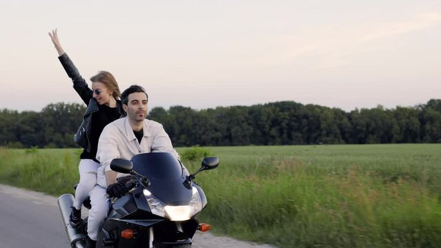 A man and a woman are riding a motorcycle on a road through a field at sunset. The girl is exhilarated, feeling a sense of freedom, and stands up, raising her hand towards the sky in joy.