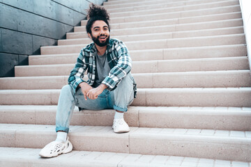 Handsome smiling hipster model. Unshaven Arabian man dressed in summer casual clothes, jeans and shirt. Fashion male with long curly hairstyle posing in the street. Sitting at the stairs