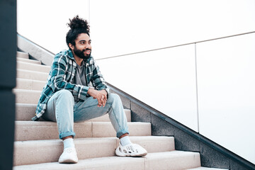 Handsome smiling hipster model. Unshaven Arabian man dressed in summer casual clothes, jeans and shirt. Fashion male with long curly hairstyle posing in the street. Sitting at the stairs - obrazy, fototapety, plakaty