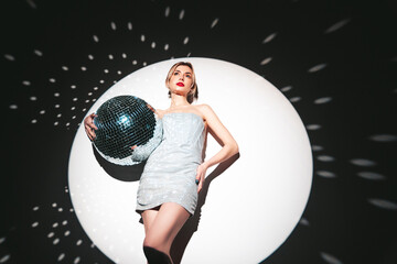 Young beautiful smiling blond female in trendy evening silver dress. Sexy carefree woman posing near white wall in studio in a circle of light. Fashionable model with bright makeup. Holds disco ball