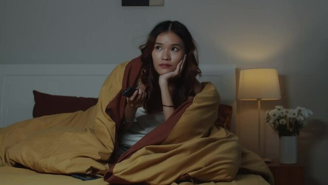 Medium shot of young girl wrapping up in blanket watching romantic comedy on TV before sleep at nighttime