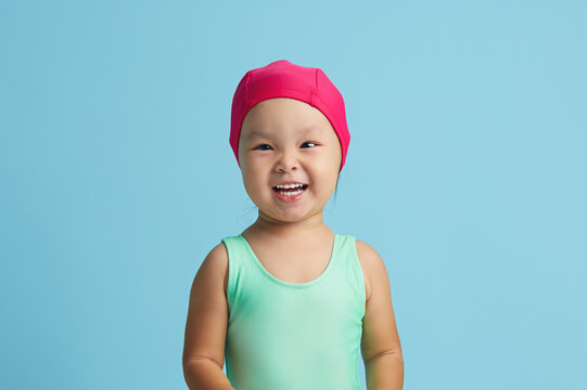 Little cheerful girl swimmer wears pink cap and swimming costume, prepares for workout in pool, smiles broadly, stands over blue background with blank space