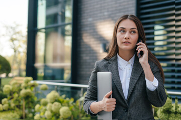 Confident business woman holding laptop and talking phone standing on modern building background....