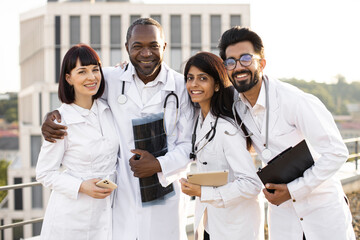 Portrait of successful team of multinational doctors in white coats with stethoscopes holding...