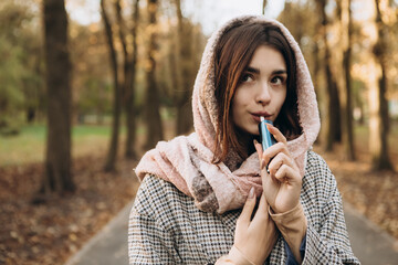 Young woman outside autumn park fall and smoking tobacco device electronic cigarette heater. Smoke...
